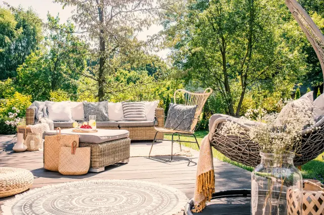 Sunny patio in a lush garden with a rattan lounge set, cushions, a low table with drinks, and decorative accessories like baskets, a throw, and flowers.